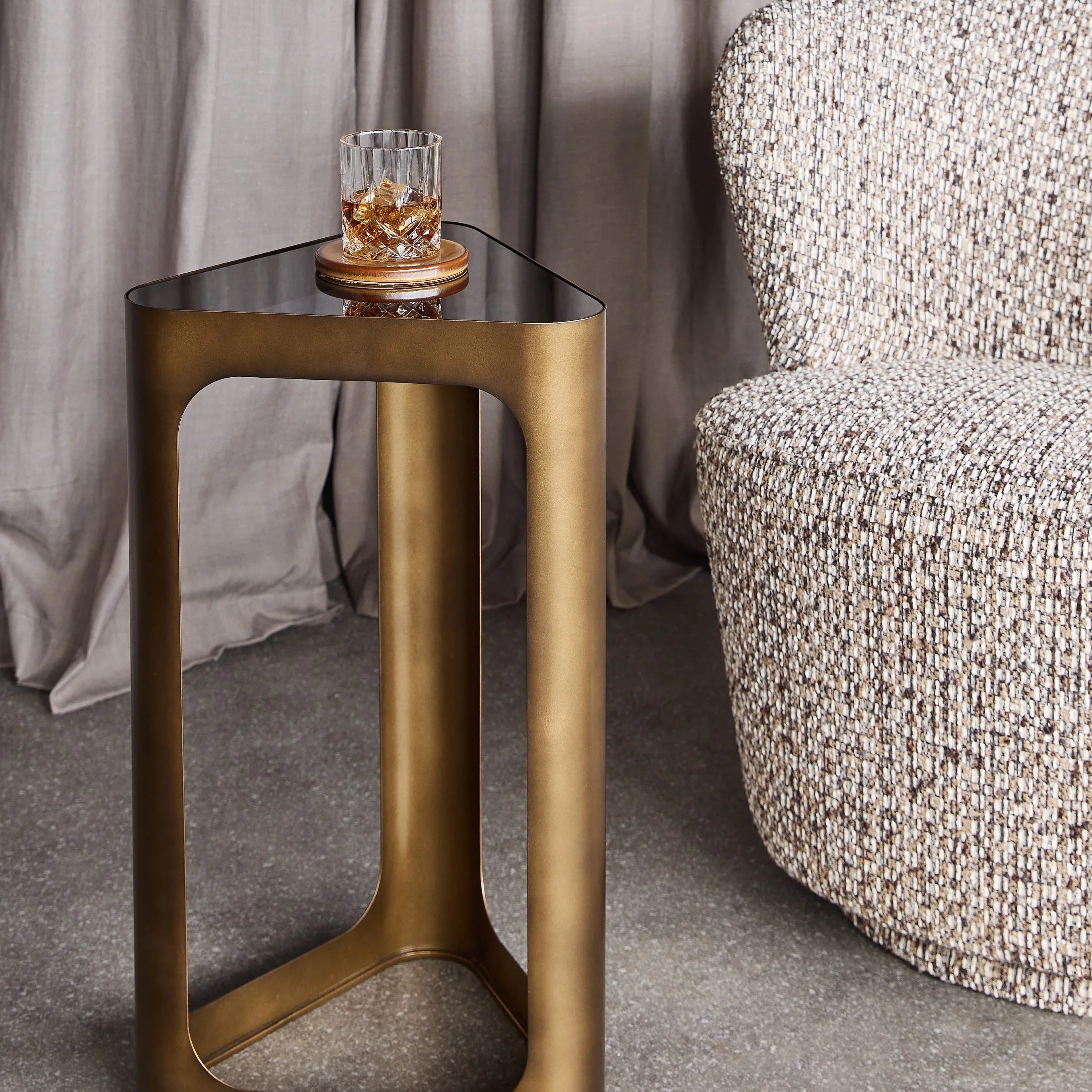 Bronze triangular table with black glass top beside a textured chair.