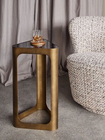 Bronze triangular table with black glass top beside a textured chair.
