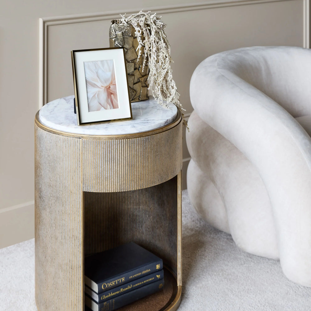 Aged gold side table with marble top next to a plush cream chair.