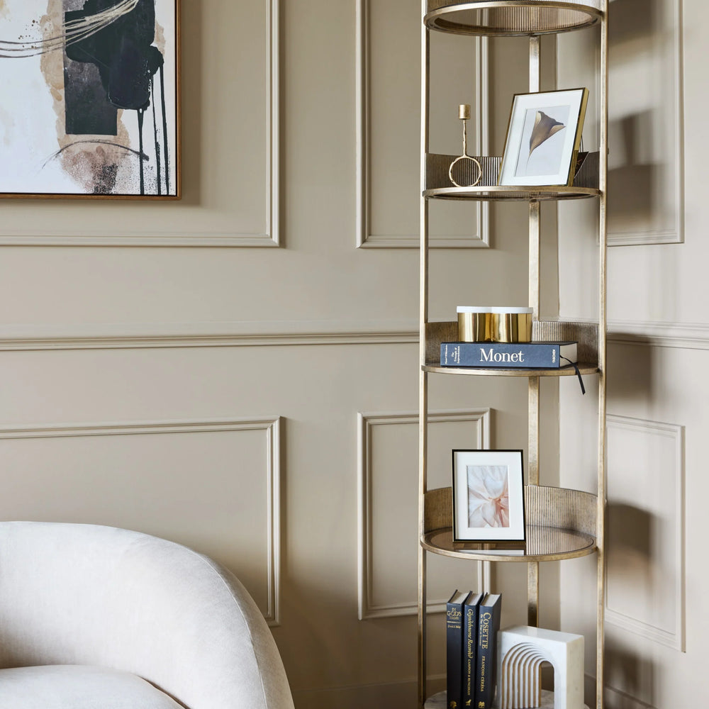 Aged gold round shelving unit in a beige room with books and decorative items.