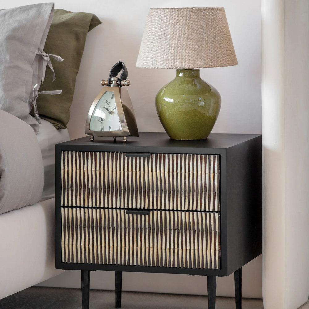 Black bedside table with bone inlay beside bed, topped with green lamp and clock.