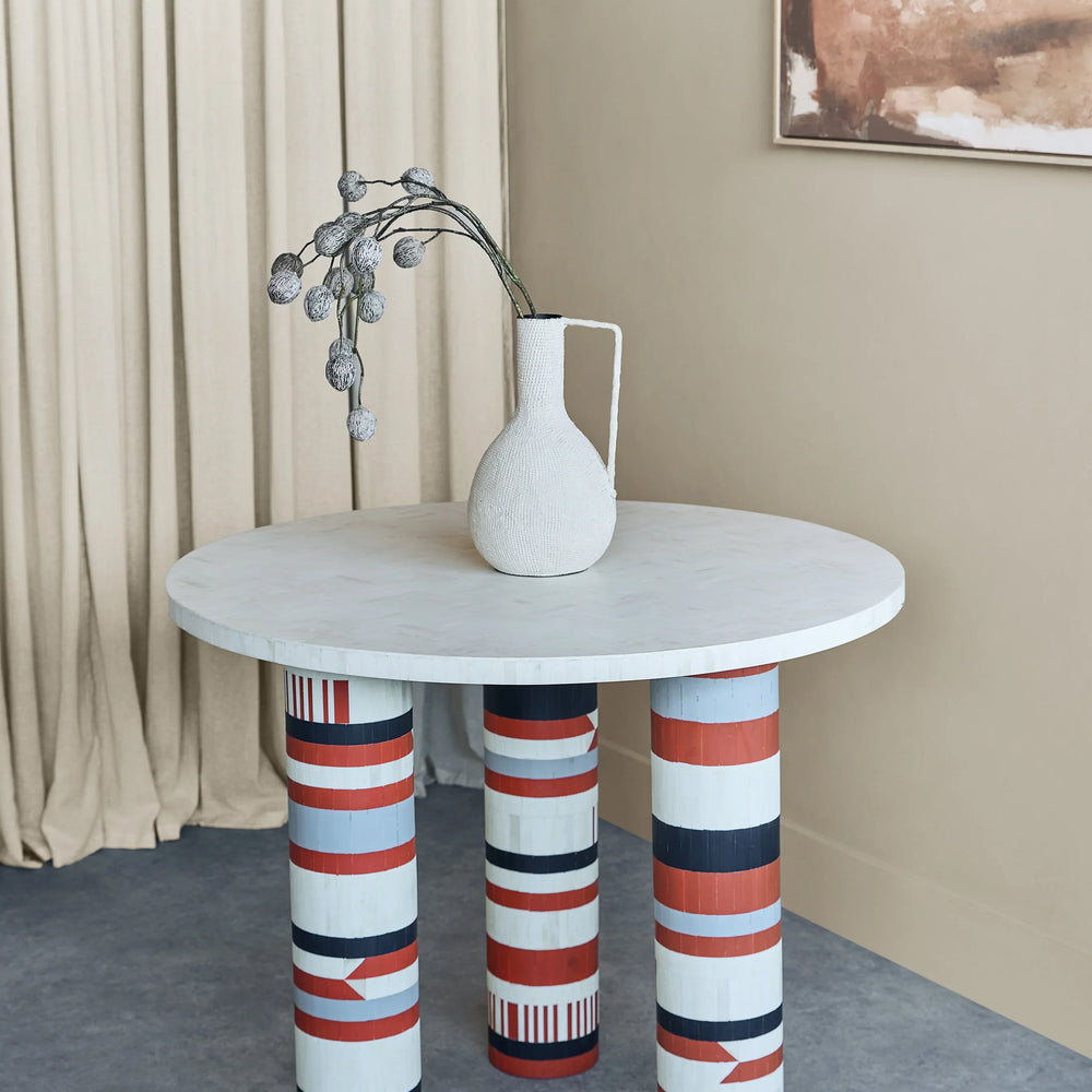 Round table with patterned legs, white vase, and dried flowers in a modern room.