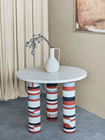 Round table with patterned legs, white vase, and dried flowers in a modern room.