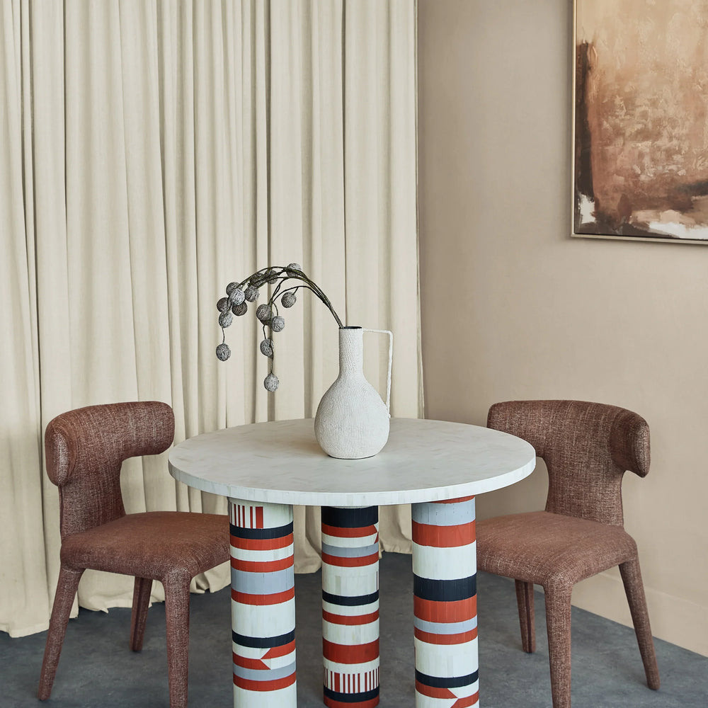 Round dining table with patterned legs, flanked by two brown chairs.