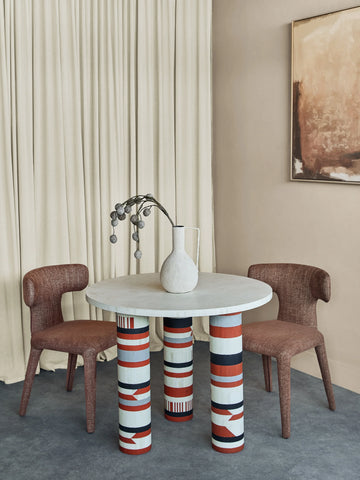 Round dining table with patterned legs, flanked by two brown chairs.