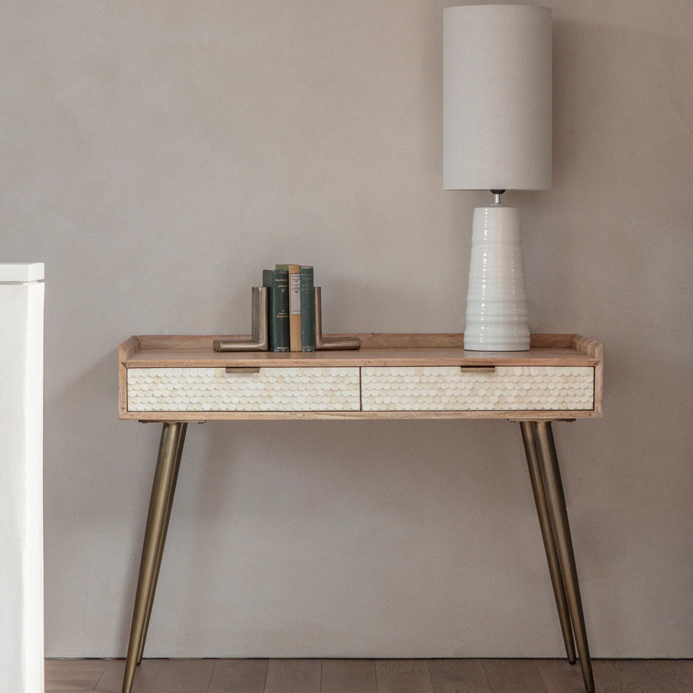 Wooden desk with bone inlay drawers, brass legs, lamp, and books in a minimalist room.