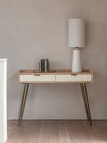 Wooden desk with bone inlay drawers, brass legs, lamp, and books in a minimalist room.