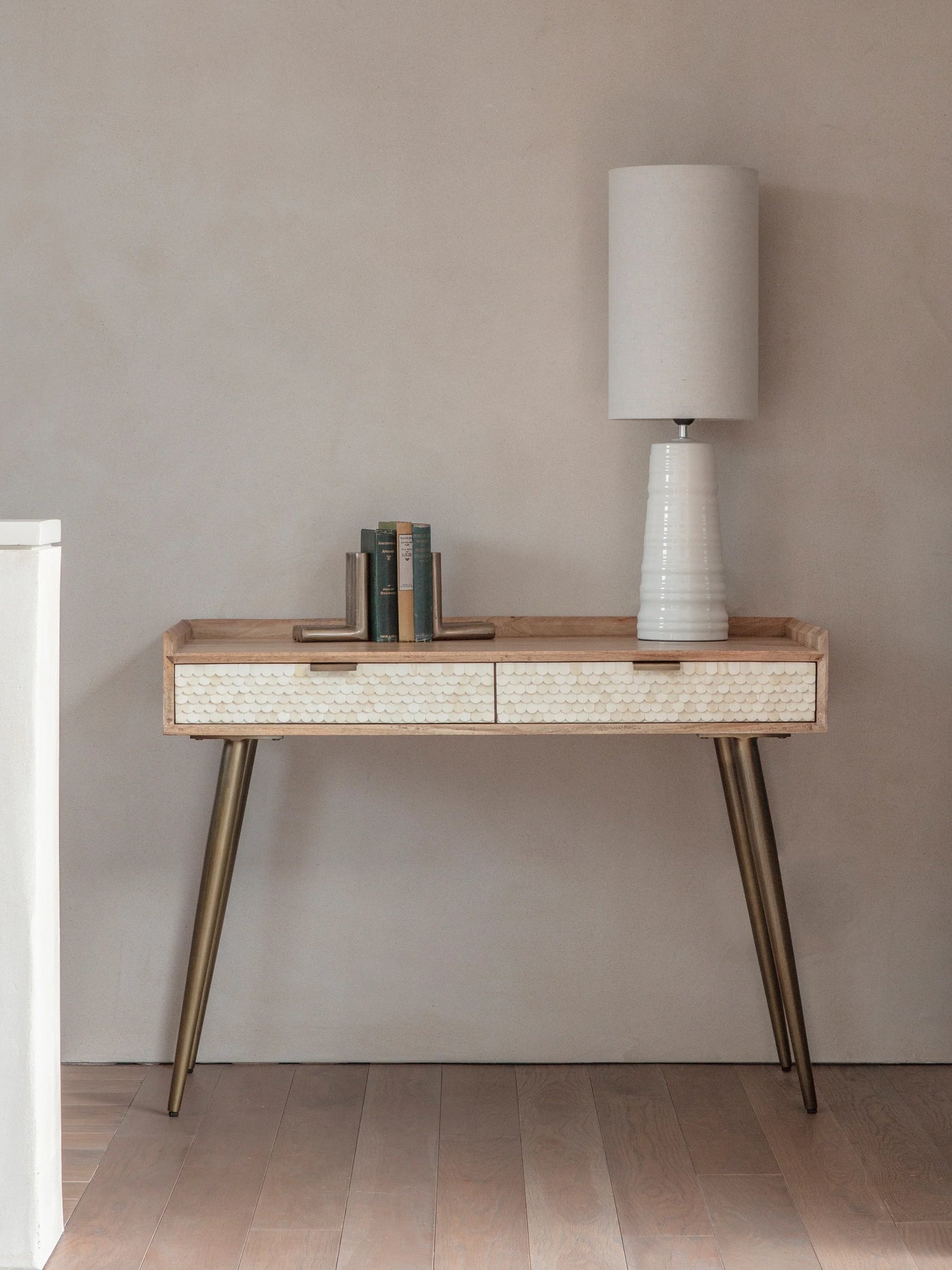 Wooden desk with bone inlay drawers, brass legs, lamp, and books in a minimalist room.
