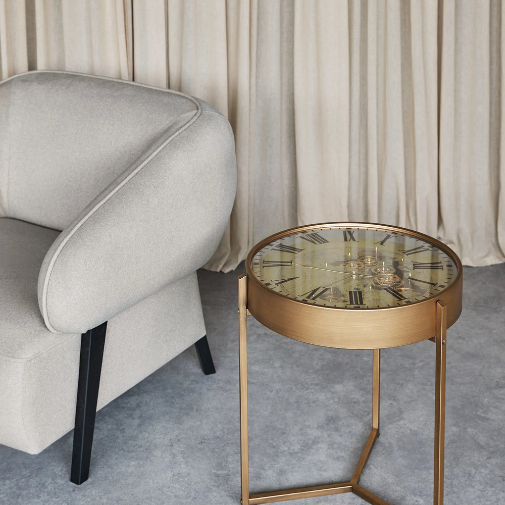 Bronze side table with clock face top beside a grey armchair on a textured floor.