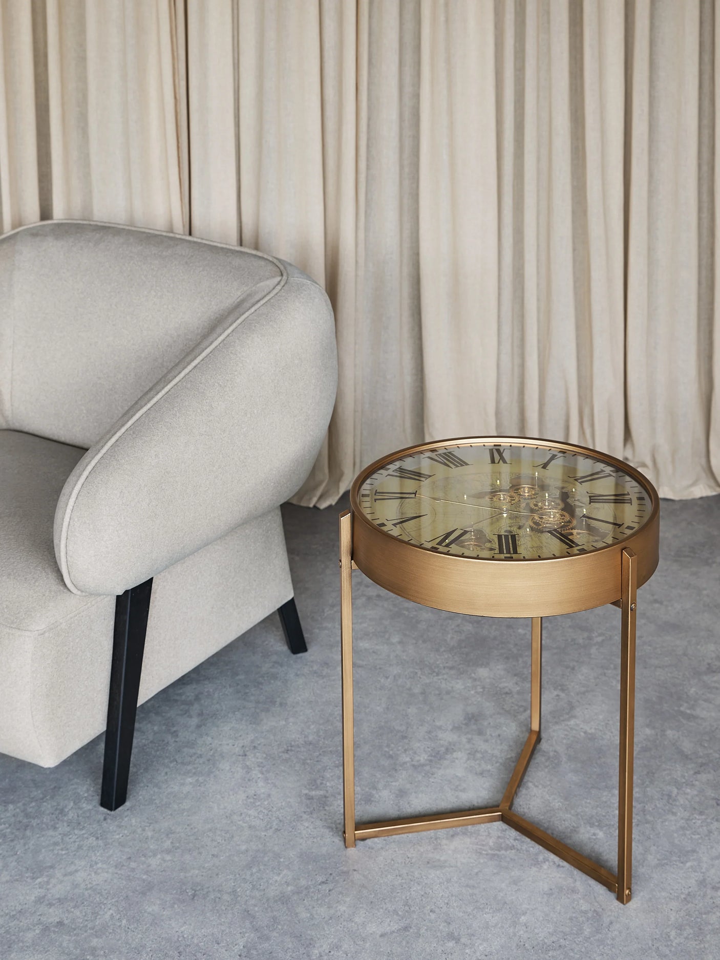 Bronze side table with clock face top beside a grey armchair on a textured floor.