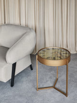 Bronze side table with clock face top beside a grey armchair on a textured floor.