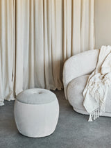 Beige storage stool in cozy living room with textured sofa and curtains.