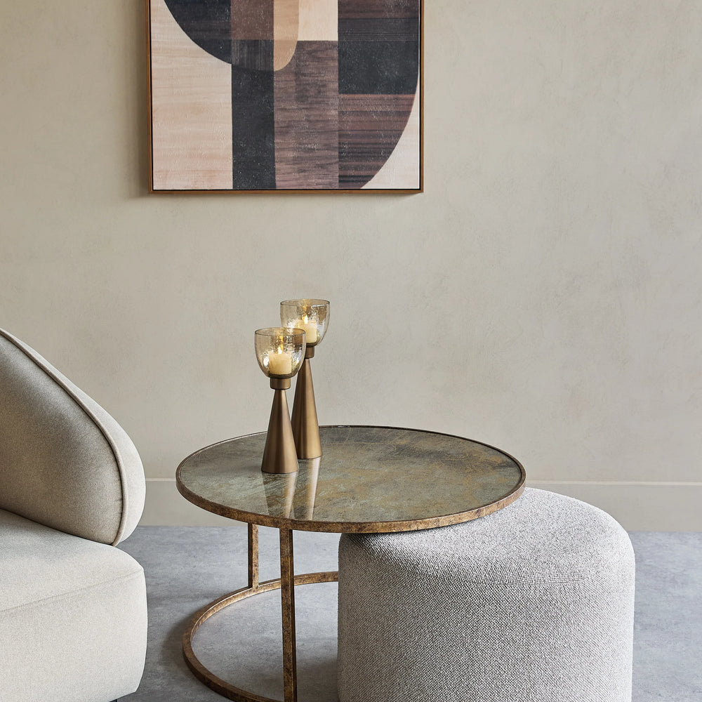Round glass coffee table with bronze base beside a textured pouffe.