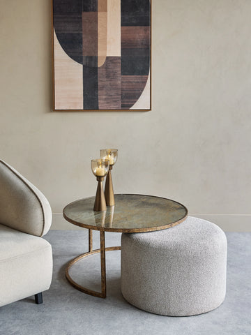 Round glass coffee table with bronze base beside a textured pouffe.