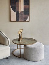 Round glass coffee table with bronze base beside a textured pouffe.