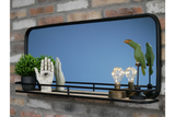 Black-framed rectangular wall mirror with wooden shelf featuring plant, decorative hands, light bulbs, and bird sculptures.