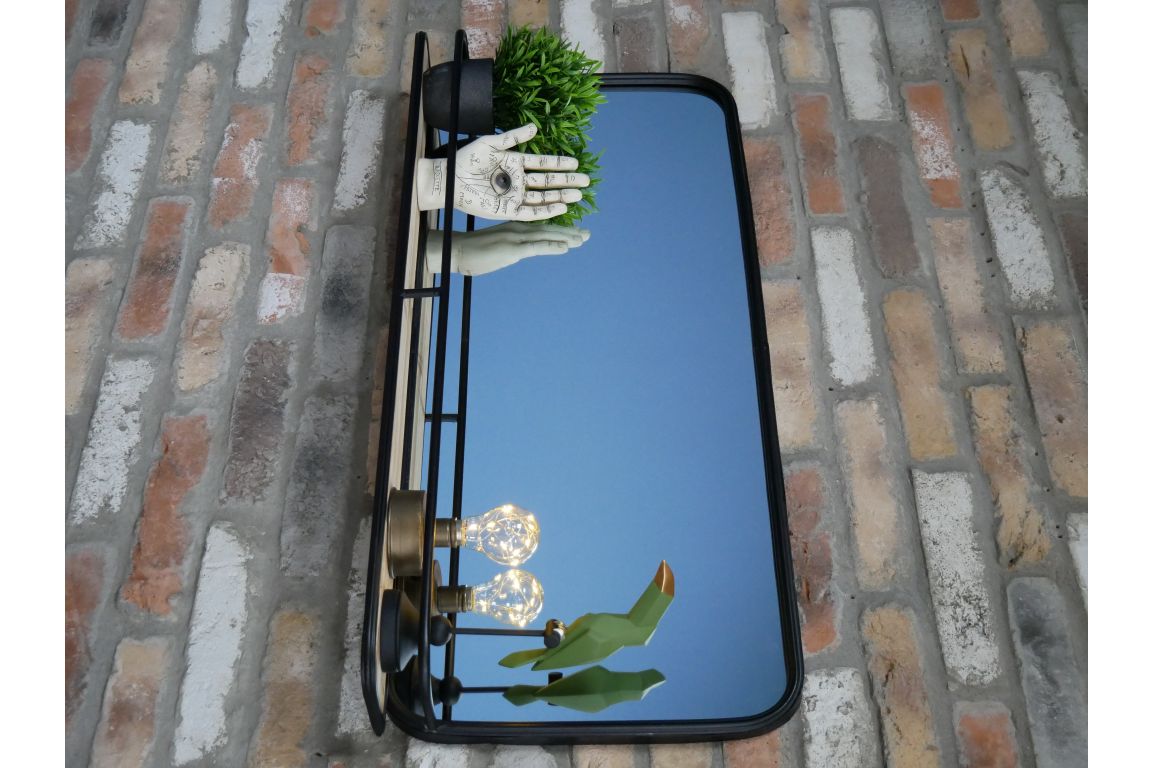 Rectangular black metal framed wall mirror with integrated shelf holding decor against a brick wall.