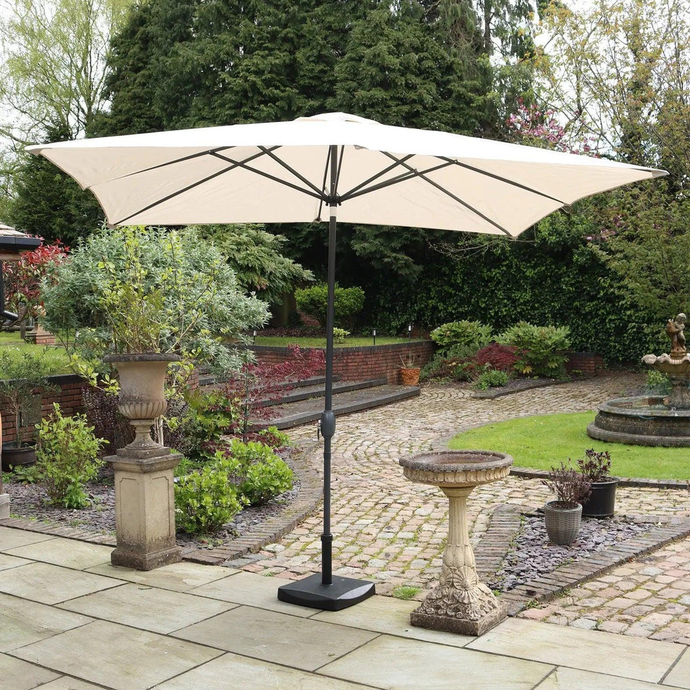 Beige garden parasol with black stand on paved patio, surrounded by greenery and stone ornaments
