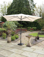 Beige garden parasol with black stand on paved patio, surrounded by greenery and stone ornaments
