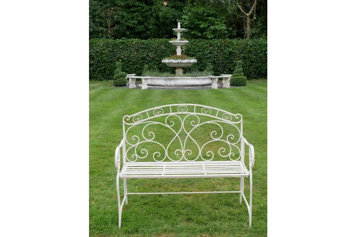 White metal garden bench with decorative scrollwork on lawn, stone fountain in background