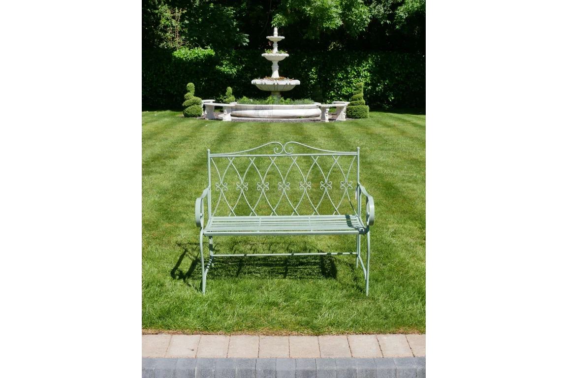 Metal garden bench with decorative backrest on lawn, fountain and topiary in background