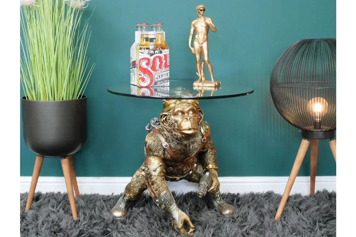 Glass side table with gold steampunk monkey base beside potted plant and modern floor lamp