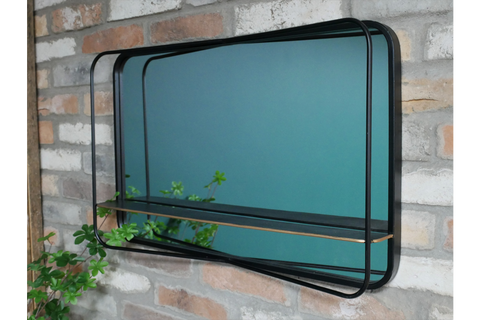 Rectangular wall mirror with black metal frame and thin shelf, angled view against brick wall with plant reflection.