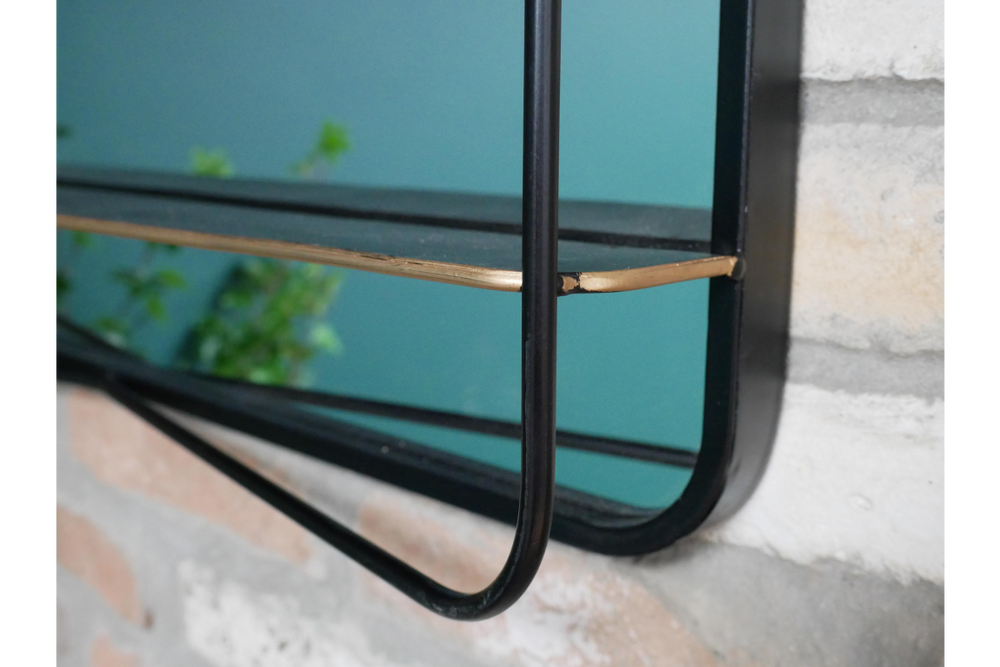 Black metal framed mirror with attached thin black shelf and gold corner detail mounted on stone wall.