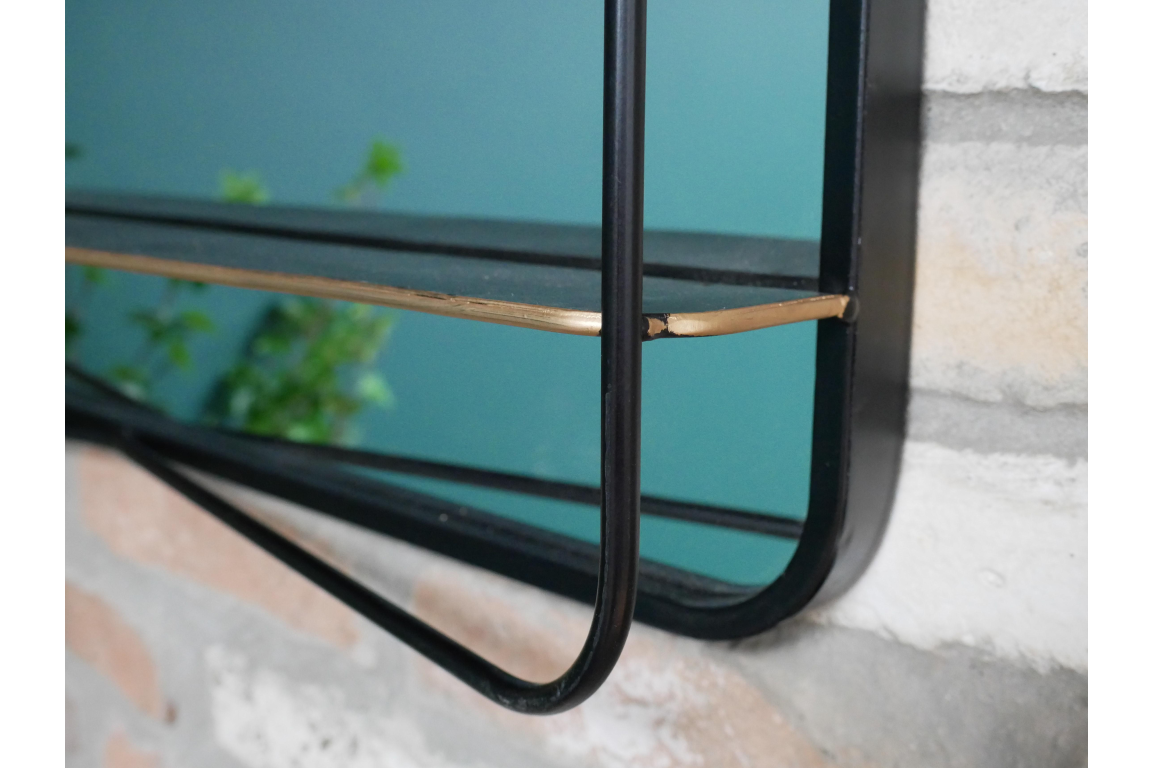Black metal framed mirror with attached thin black shelf and gold corner detail mounted on stone wall.