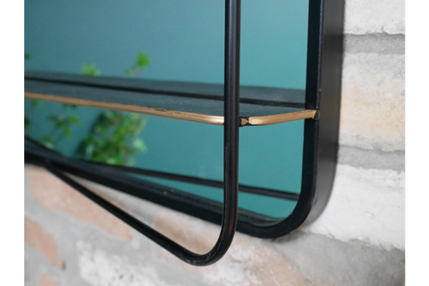 Black metal framed mirror with attached thin black shelf and gold corner detail mounted on stone wall.