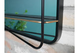 Black metal framed mirror with attached thin black shelf and gold corner detail mounted on stone wall.