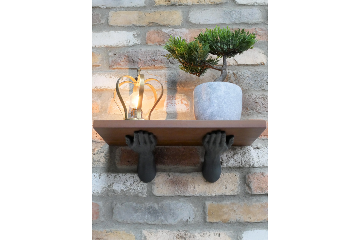 Wooden wall shelf held by two black metal hand brackets, displaying a potted plant and decorative light, brick wall