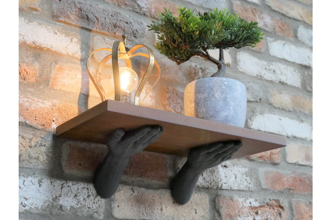 Wall-mounted wooden shelf with black sculpted hands as support, shown with a plant pot and decorative lamp.