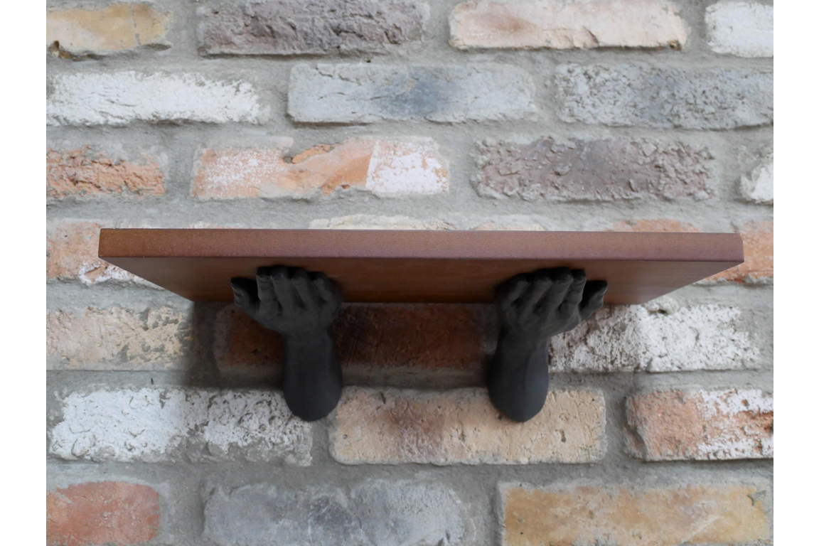 Wall-mounted wooden shelf held by two black hands against a rustic brick wall, front view.