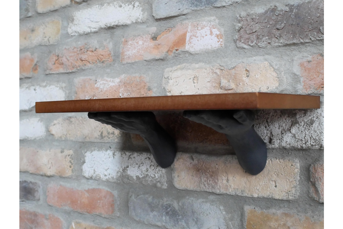 Wall-mounted brown wooden shelf supported by two black hands, angled view against a brick wall background.