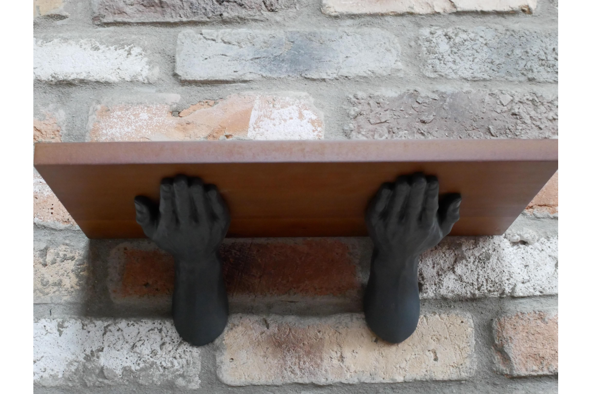 Wall-mounted wooden shelf held by two black sculpted hands against a rustic brick wall backdrop.