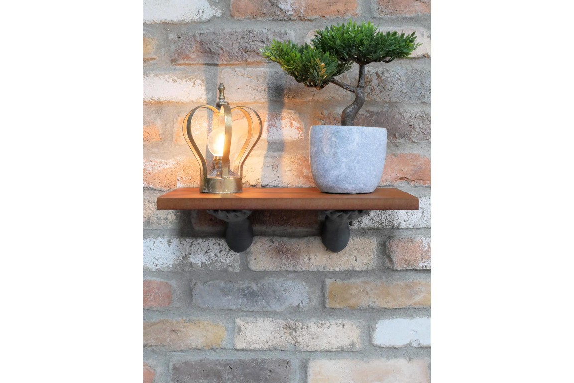 Wall-mounted rectangular wooden shelf with two black hand-shaped brackets holding a lamp and a potted plant, brick