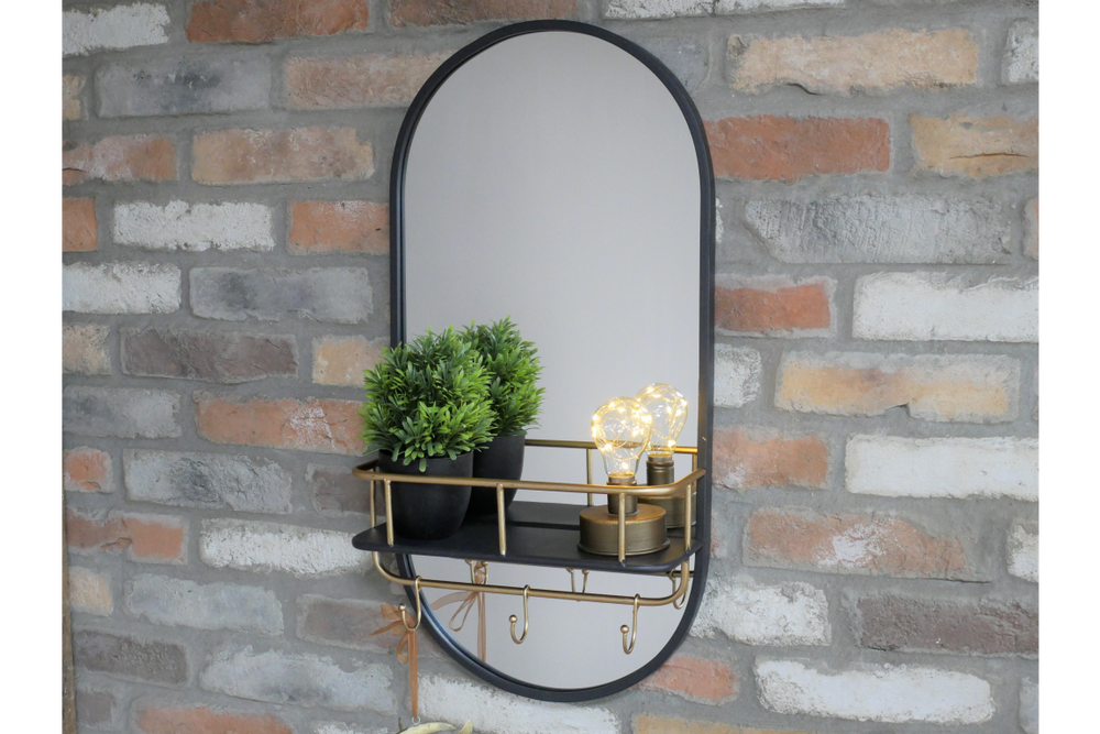 Oval black-framed wall mirror with black and brass shelf holding two plants and two light bulbs, mounted on brick wall.