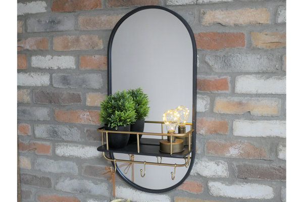 Oval black-framed wall mirror with black and brass shelf holding two plants and two light bulbs, mounted on brick wall.