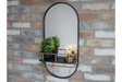 Oval black-framed wall mirror with black and brass shelf holding two plants and two light bulbs, mounted on brick wall.