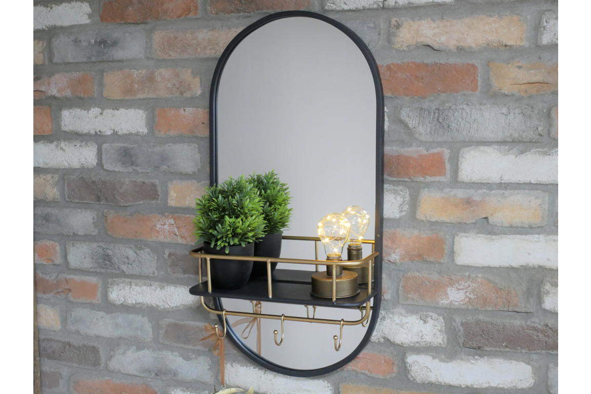 Oval black-framed wall mirror with black and brass shelf holding two plants and two light bulbs, mounted on brick wall.