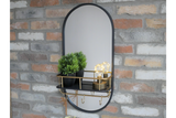 Oval black-framed wall mirror with black and brass shelf holding two plants and two light bulbs, mounted on brick wall.