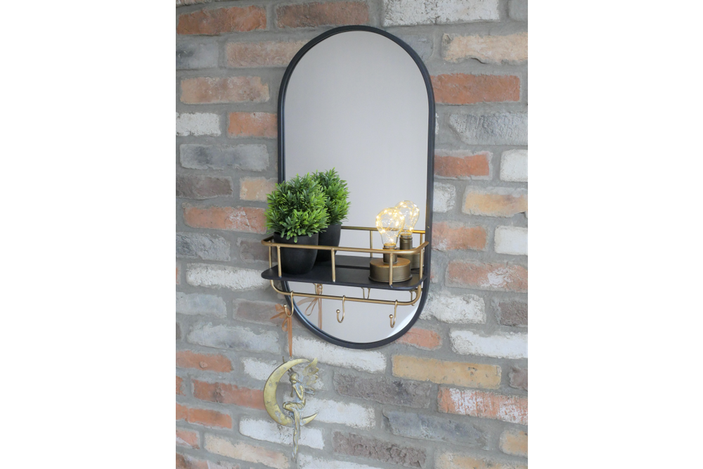 Oval wall-mounted mirror with black frame, gold metal shelf and hooks, shown with potted plant and decorative bulbs.