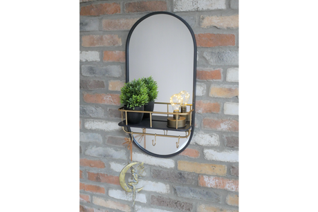 Oval wall-mounted mirror with black frame, gold metal shelf and hooks, shown with potted plant and decorative bulbs.