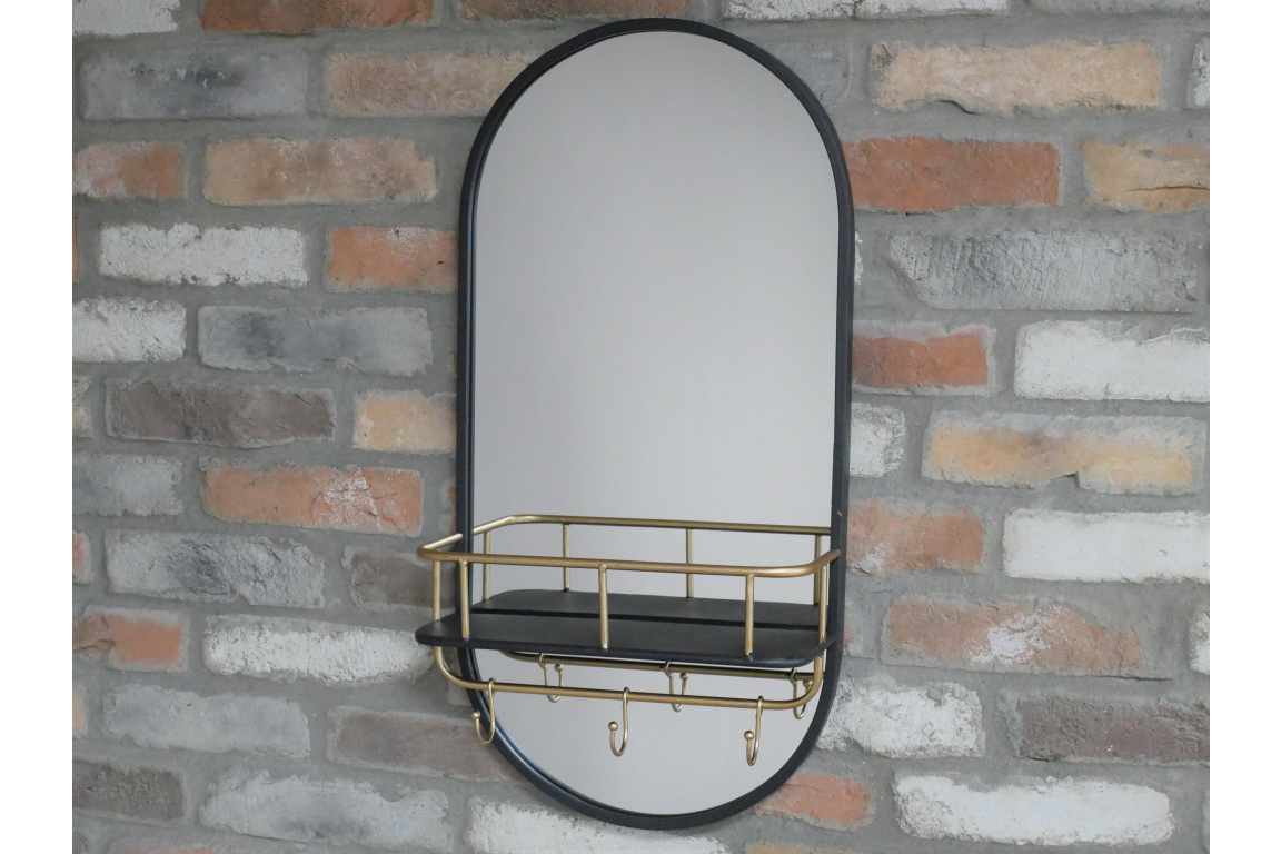 Oval wall mirror with black frame, attached black shelf with gold railing and hooks, mounted on brick wall.
