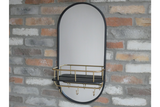 Oval wall mirror with black frame, attached black shelf with gold railing and hooks, mounted on brick wall.