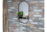 Oval wall mirror with black frame and brass shelf with hooks, decorated with two small plants and light bulbs.