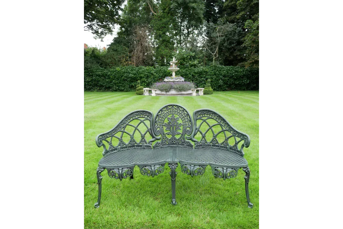 Cast iron garden bench with ornate backrest on lawn, fountain and trees in background