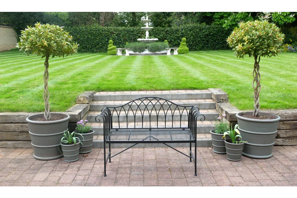 Black metal garden bench on patio with potted plants, facing striped lawn and stone steps