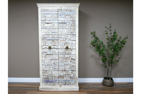 Large white wooden cabinet with intricate carved details and metal ring handles, standing on wooden floor beside green plant.